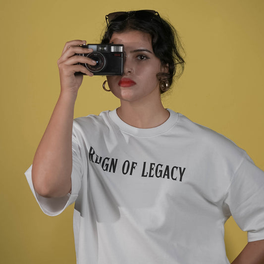 Person wearing a white t-shirt with ' Reign of Legacy ' text, holding a camera against a yellow background